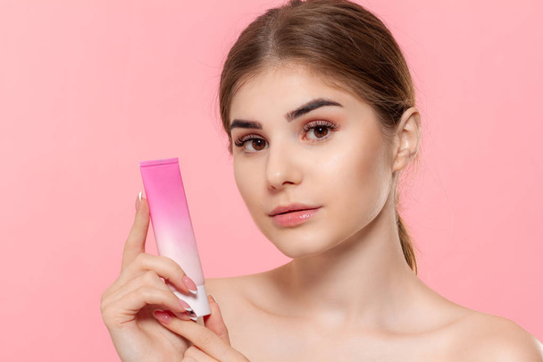 Linda menina segurando tubo de creme para hidratar a pele. Modelo olha para a câmera isolada sobre fundo rosa
. - Foto, Imagem