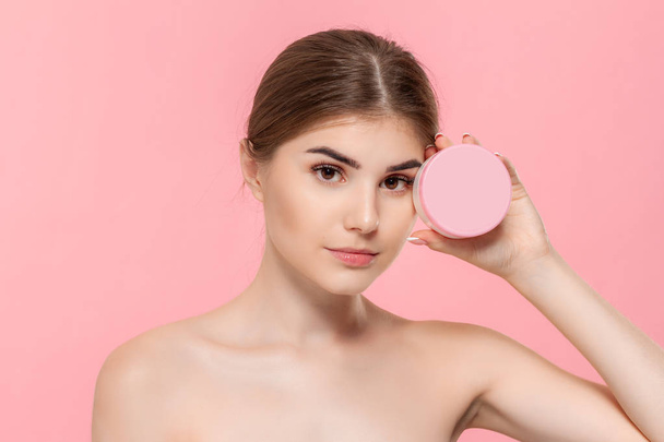 Beautiful girl holding tube of cream to moisturize skin. Model looks at the camera isolated over pink background. Concept of beauty and health treatment. - Photo, Image