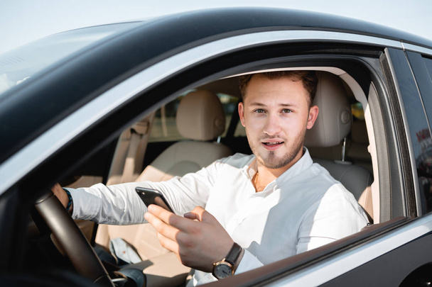 Fiatal üzletember mosolyogva nézi mobiltelefonján vezetés közben egy autó. - Fotó, kép