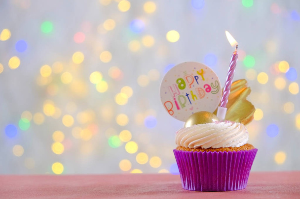 süßer Bio-Happy-Birthday-Cupcake mit brennender Kerze in lila Verpackung, Frischkäse-Zuckerguss-Strudel und goldener Schokoladendekoration auf weißem Holztisch. Nahaufnahme, Kopierraum, Hintergrund. - Foto, Bild