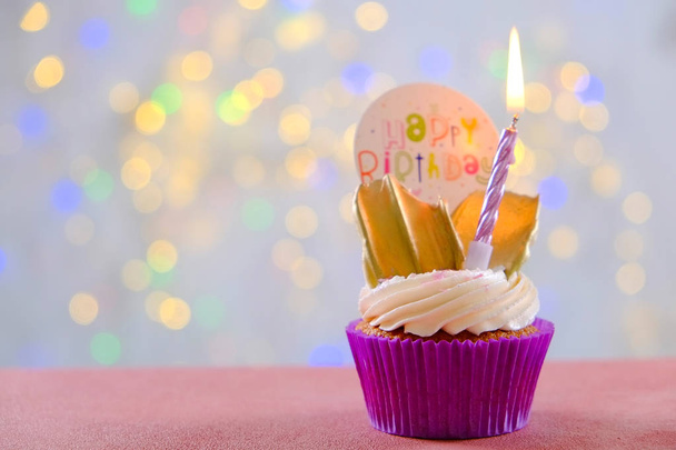 Sweet organic happy birthday cupcake with burning candle in purple wrap, cream cheese frosting swirl and golden chocolate decoration on white wooden textured table. Close up, copy space, background. - Photo, Image