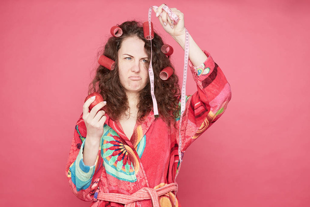Depressive attractive emotional woman looks with disappointment at measuring tape, holds fresh red apple in hand, dressed in home colorful  bathrobe, isolates on pink studio wall with blank copy space. - Fotó, kép