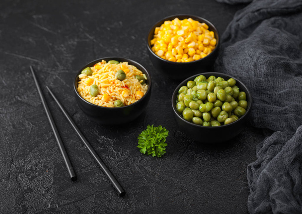 Black bowls with boiled organic basmati vegetable rice, yellow corn and peas with black chopsticks on black stone background with linen cloth. Top view - Foto, Bild