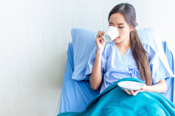 Ασιατικές νεαρές γυναίκες ασθενείς πίνουν ζεστό νερό σε ένα κρεβάτι ασθενούς στο δωμάτιο ανάρρωσης. Και φωτεινές εκφράσεις του προσώπου στον θάλαμο του ασθενούς. - Φωτογραφία, εικόνα