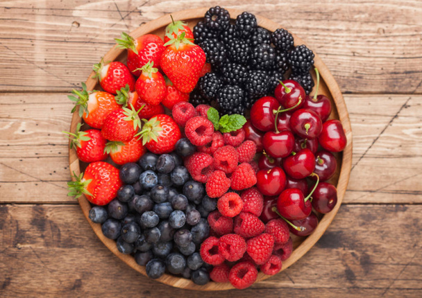 Fresh organic summer berries mix in round wooden tray on light wooden table background. Raspberries, strawberries, blueberries, blackberries and cherries. - Photo, Image