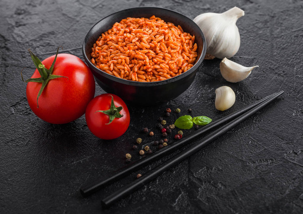 Black plate bowl of rice with tomato and basil and garlic and chopsticks on black stone background. Traditional food. - Фото, зображення