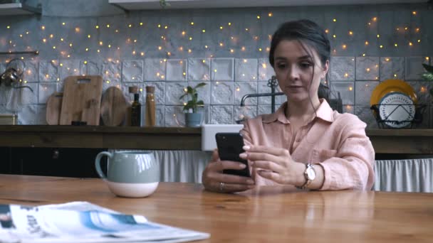 Close-up view of pretty young woman with brown hair in pink shirt sitting in the kitchen at wooden table and using smartphone. Stock footage. Modern decorated home interior on the background - Video, Çekim