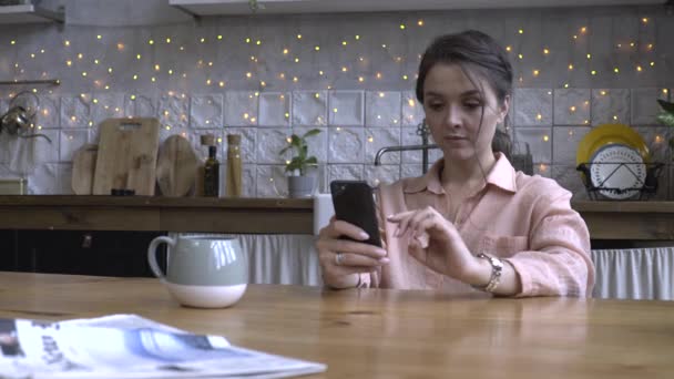 Beautiful young woman with brown hair in pink shirt sitting in the kitchen at wooden table and using smartphone. Stock footage. Modern decorated home interior on the background - Metraje, vídeo