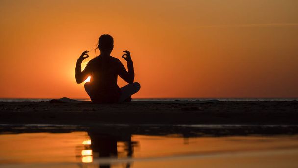 Młoda kobieta siedzi nad morzem w pozycji lotosu o zachodzie słońca, medytuje - Zdjęcie, obraz