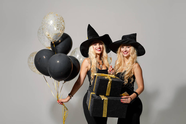 Two happy young women in black witch halloween costumes holding a glass of champagne and gift boxes on party over white background. Sale and gift concept. - Foto, imagen