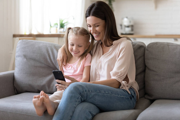 Positive mother embraces daughter sitting on couch using smartphone - Foto, Bild