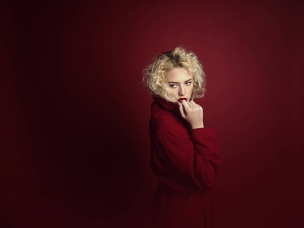 Piękny portret uwodzicielski kobieta z jasnym blond włosy, niebieskie oczy i czerwone usta stwarzające w czerwonym płaszczu i na ciemnoczerwonym tle. Moda, glamour, koncepcja - Zdjęcie, obraz