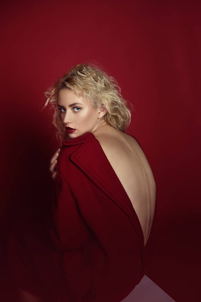 Beautiful portrait seductive woman with light blond hair,blue eyes and red lips posing in red coat and on dark red background. Fashion, glamour, concept - Фото, изображение