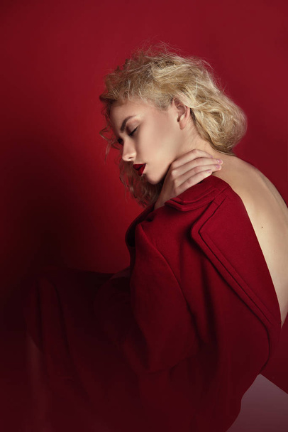 Hermosa mujer seductora retrato con pelo rubio claro, ojos azules y labios rojos posando en abrigo rojo y sobre fondo rojo oscuro. Moda, glamour, concepto
 - Foto, imagen