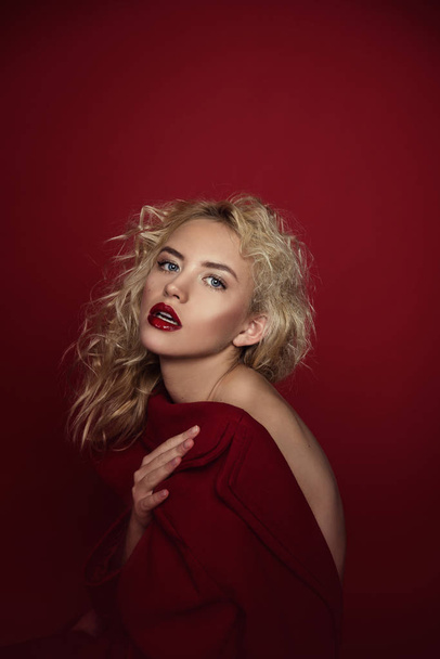 Beautiful portrait seductive woman with light blond hair,blue eyes and red lips posing in red coat and on dark red background. Fashion, glamour, concept - Fotó, kép