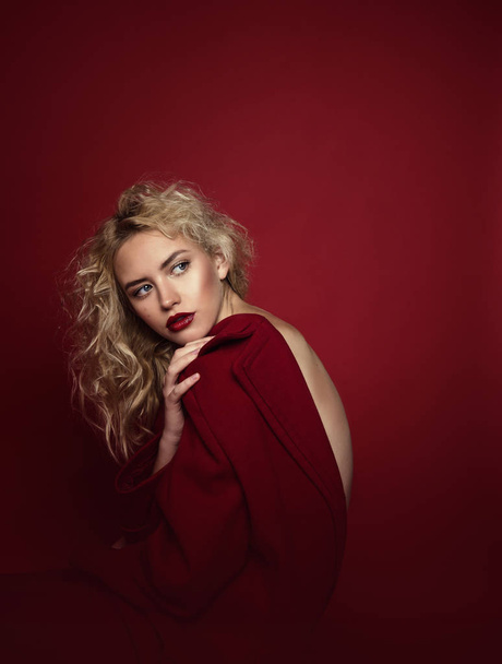 Beautiful portrait seductive woman with light blond hair,blue eyes and red lips posing in red coat and on dark red background. Fashion, glamour, concept - Foto, imagen
