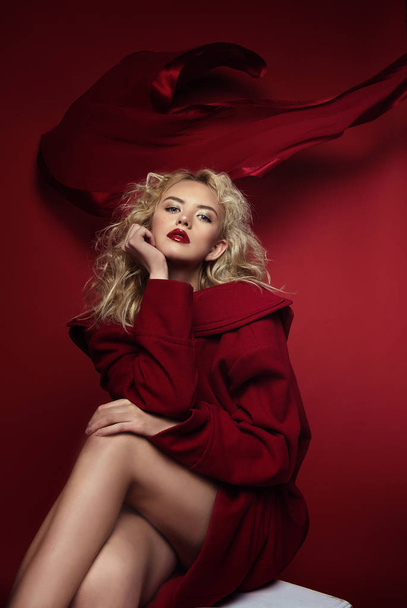Beautiful portrait seductive woman with light blond hair,blue eyes and red lips posing in red coat and on dark red background. Fashion, glamour, concept - Foto, Imagem