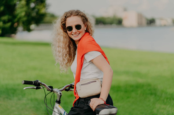 glücklich gelockte blonde Frau genießt Fahrradfahren, hat ihren eigenen Transport, führt einen gesunden Lebensstil, fährt mit dem Fahrrad durch malerische Naturlandschaften, trägt legeres Outfit, hat aktiven Urlaub - Foto, Bild