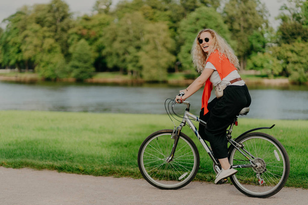 Outdoor shot van gelukkige blonde krullende vrouw draagt een zonnebril, gekleed in casual actieve draagt, ritten op de fiets in de buurt van het meer en groene bomen op het platteland, brengt vrije tijd buiten, geniet van favoriete hobby - Foto, afbeelding