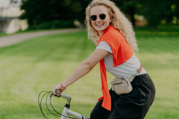 Vízszintes lövés boldog göndör nő lovagol kerékpár, tartja a kezét a kormányon, úgy néz ki, aktív és frissített, öltözött alkalmi, visel árnyalatok, pózol a zöld természet háttér. Spare time koncepció - Fotó, kép