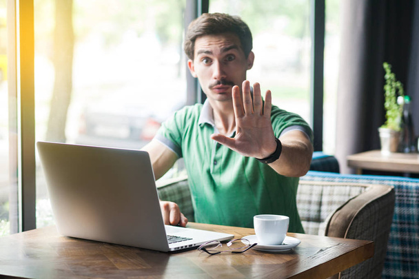 Young serious businessman in green t-shirt sitting with laptop in cafe and looking at camera while showing stop sign gesture. business and freelancing concept  - 写真・画像