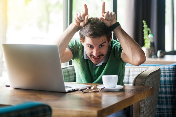 Young angry businessman in green t-shirt sitting with laptop in cafe and looking at camera with crazy face and showing cow horns gesture on head. business and freelancing concept. - Fotó, kép
