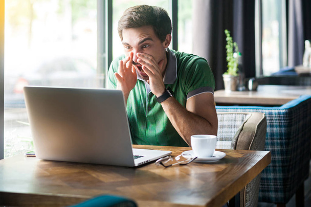 junger Geschäftsmann in grünem T-Shirt sitzt im Café und schaut auf Laptop-Display während Videoanruf und schreit  - Foto, Bild