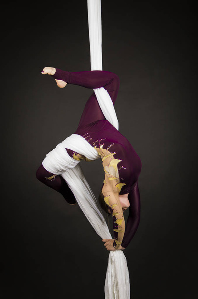 Sporty girl in a Burgundy suit performs gymnastic and circus exercises on white silk. - Фото, изображение