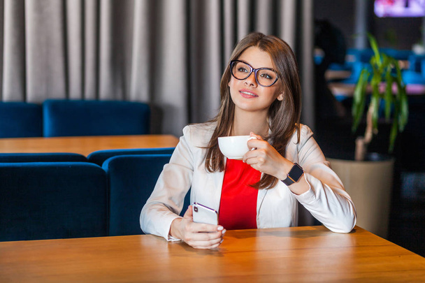 joyeuse belle brune élégante jeune femme dans des lunettes tenant une tasse de café et se reposant tout en étant assis à la table dans un café
  - Photo, image