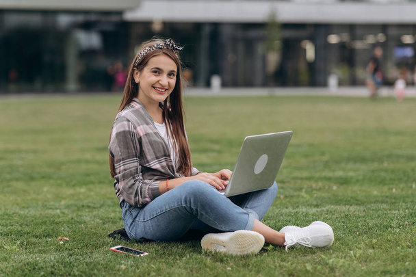 Pretty young woman sitting on green grass in park with legs crossed during summer day while using laptop - Foto, afbeelding