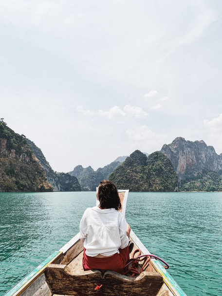 Mujer joven con falda roja y blusa blanca sentada en un barco de madera observando islas grandes exóticas y tropicales de color verde oscuro con rocas y lago turquesa en el lago Cheow Lan, Khao Phang, distrito de Ban Ta Khun, Tailandia. Viajes vacaciones y concepto de aventura
. - Foto, Imagen