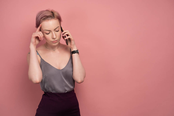 A girl with short pink hair and a tattoo in a gray top and speaks on the phone, listening carefully, on a pink background. - Foto, Bild