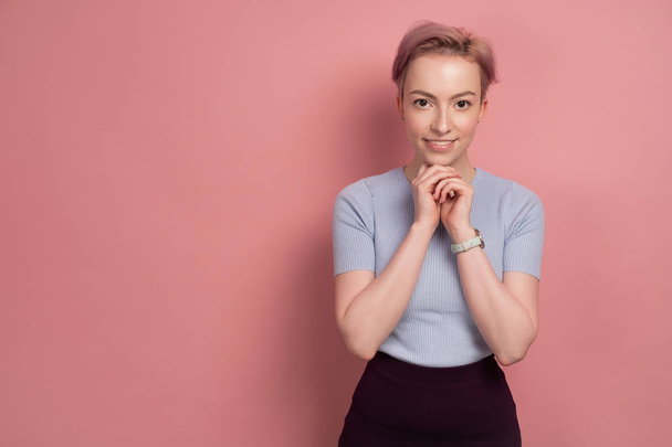 A girl with short pink hair in a T-shirt stands on a pink background, looking at the camera and propping her chin with her hands. - Foto, afbeelding
