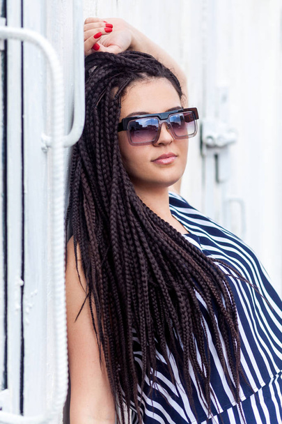 young brunette woman with dreadlocks hairstyle in striped dress and sunglasses posing and looking at camera while leaning at wall outdoor   - Fotografie, Obrázek