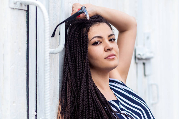 young brunette woman with dreadlocks hairstyle in striped dress holding sunglasses and looking at camera while leaning at wall outdoor   - Photo, image