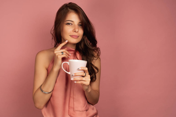 A dark-haired girl in a pink dress with a mug in her hand stands on a pink background and looks at the camera with squinted eyes. - Photo, Image