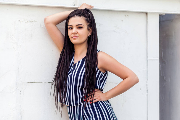 young brunette woman with black afro american dreadlocks hairstyle in striped dress posing and looking at camera while smiling   near wall at street  - Photo, image