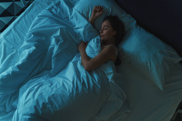 Young beautiful girl or woman sleeping alone in big bed at night, top view, blue toned - Foto, Bild