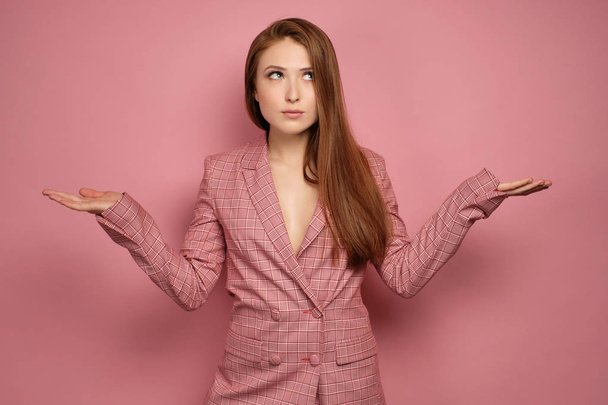 A red-haired girl in a pink jacket stands on a pink background spreading her arms to the side and looking up thoughtfully. - Foto, Imagem