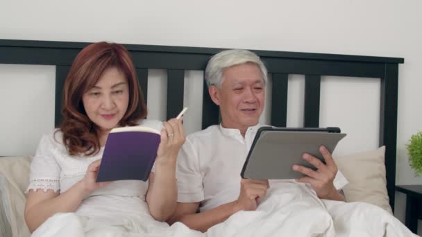 Asian senior couple using tablet at home. Asian Senior Chinese grandparents, husband watch movie and wife read book after wake up, lying on bed in bedroom at home in the morning concept. Slow motion. - Video, Çekim