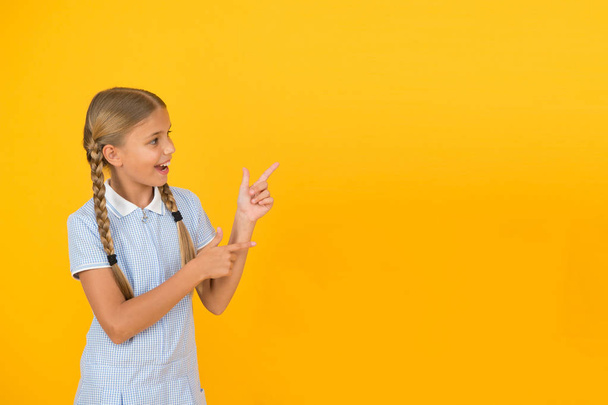 Schauen Sie sich das an. fröhliche kleine Schulmädchen zeigt mit dem Finger auf gelben Hintergrund Kopierraum. zurück zur Schule. Aufmerksamkeitskonzept. Werbemodell. Schülerin präsentiert Schulprodukt. Positives Kind - Foto, Bild