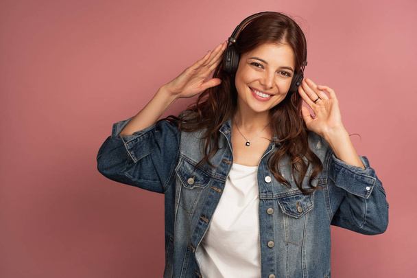 A brunette on a pink background in a denim shirt is smiling and looking into the camera while holding the headphones in her hands - Fotoğraf, Görsel