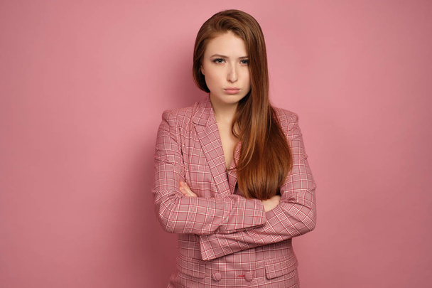 A redhead girl in a pink jacket stands on a pink background with her arms folded in front of her and looks offended at the camera - Foto, Bild