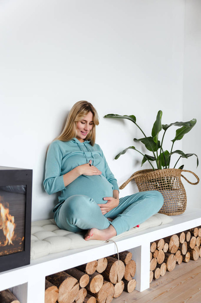 Pregnant girl in green sportswear hugs the unborn baby and practices yoga, in cozy interior - Photo, Image