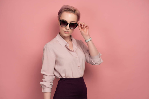 A girl with short pink hair in a blouse and skirt stands on a pink background, adjusting her sunglasses. - Foto, immagini