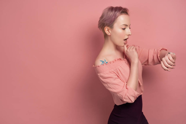 A girl with short pink hair is standing in profile in a blouse on a pink background and opening her mouth looks at the clock. - Foto, immagini