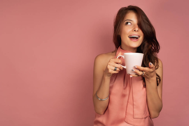 A dark-haired girl in a pink dress with a mug in her hand stands on a pink background and, opening her mouth, looks up. - Foto, Imagen
