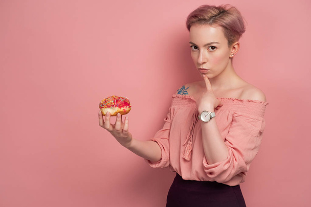A girl with short pink hair in a blouse looks thoughtfully at the camera, holding a donut in her hand, on a pink background. - Foto, Imagen