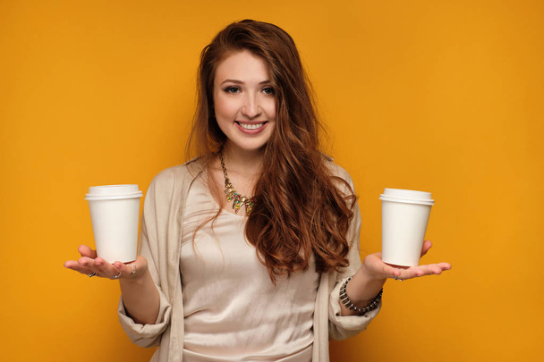 Red-haired girl stands on a yellow background smiling looking at the camera and holding two cups on the palms. - Foto, Bild