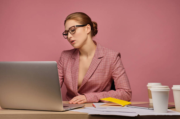 Punatukkainen tyttö, jolla on takki ja lasit, istuu pöydässä papereiden kanssa ja katselee väsymättä kannettavaa tietokonetta
. - Valokuva, kuva